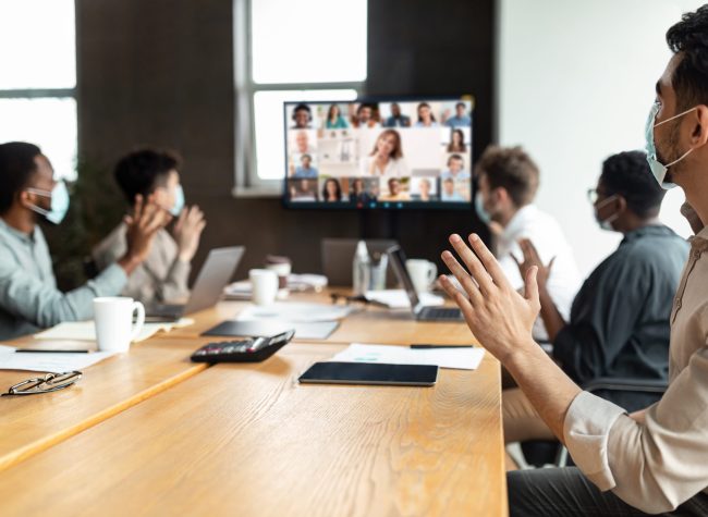 Videoconferencing Concept. Diverse group of workers in face masks making video call with colleagues, sitting at table in boardroom, waving at screen at office, talking on web with remote employees